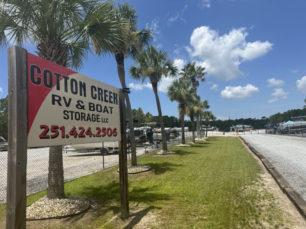 Cotton Creek RV & Boat Storage LLC sign showing phone 251.424.2506, palm trees, and storage lot in background.